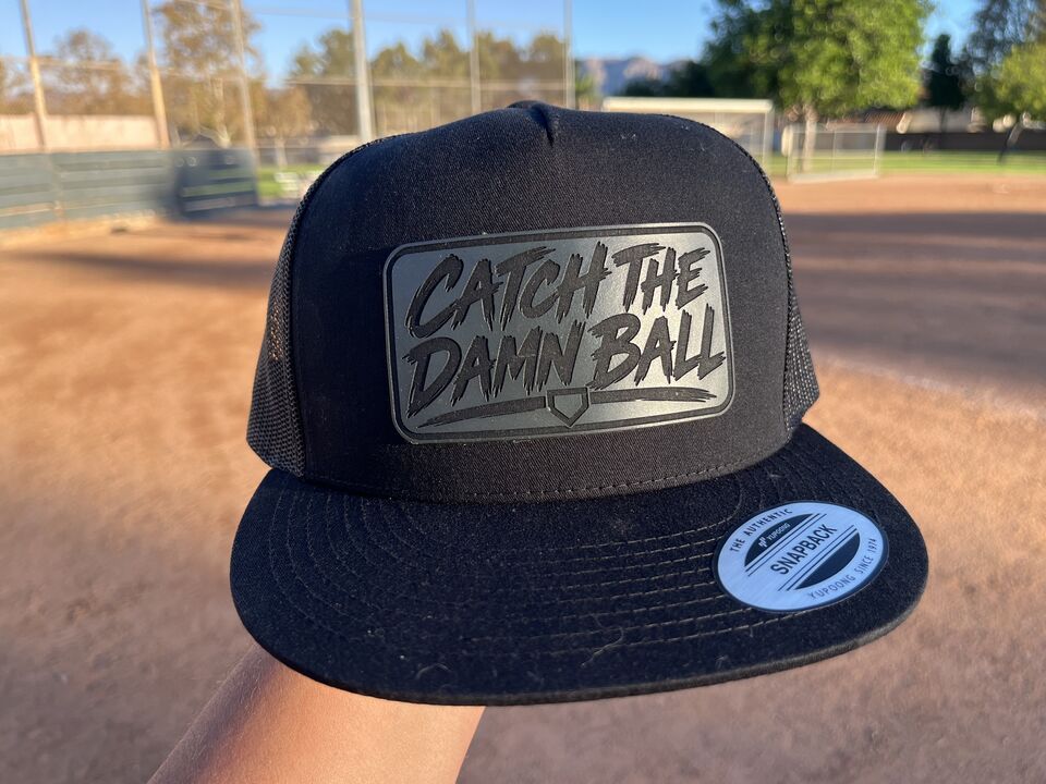 The black mesh back Catch The Damn Ball patch hat held up at the ball field