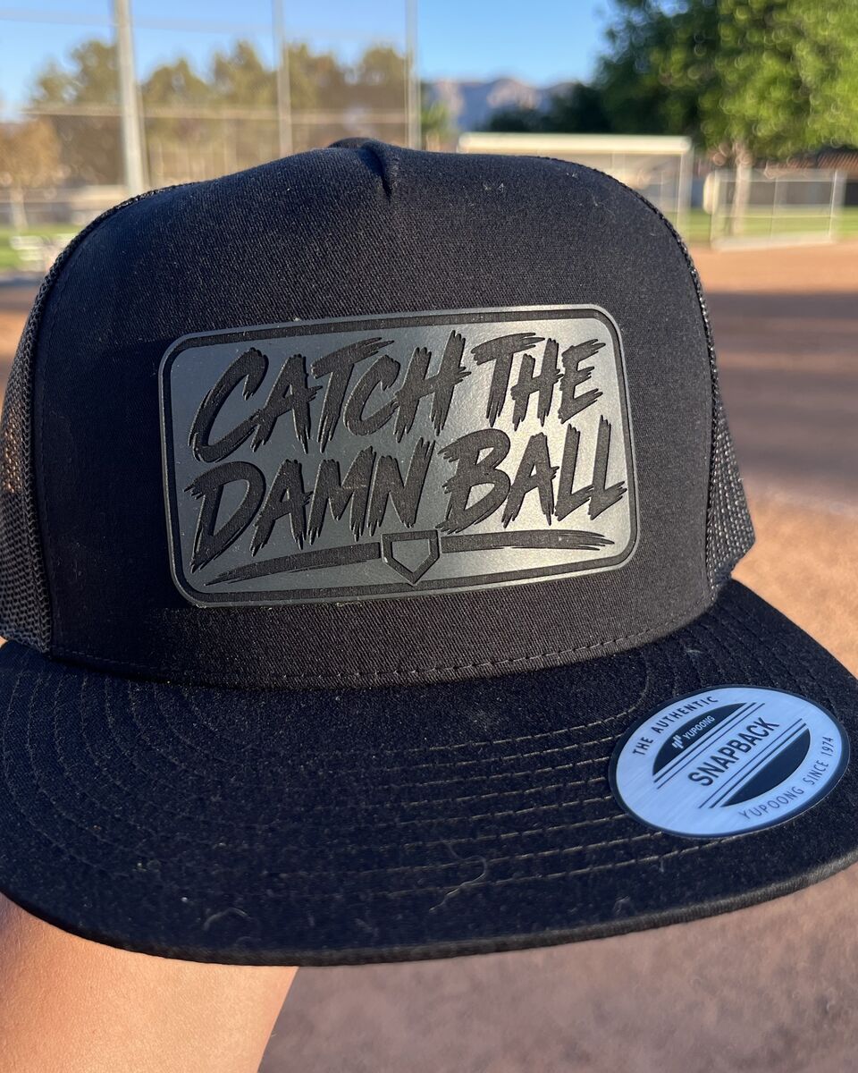 Black mesh back hat with the Catch The Damn Ball patch, held up at the ball field